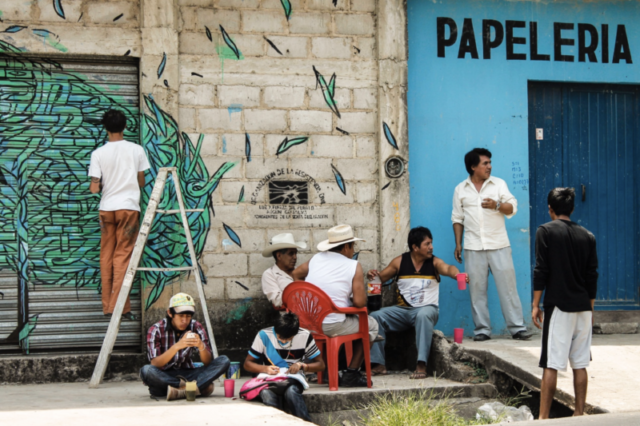 A group of activists sitting in front of one of their houses, while street-artist Crisa paints a mural. The seal of Luz y Fuerza del Pueblo appears on the wall behind them. - Comalapa, Chiapas. April 2014.